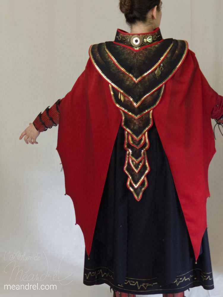 Une photo de dos d'un costume composé d'une cape noire longue, sous une cape rouge ouverte en forme d'ailes triangulaires, couronnées d'un plastron noir à bords ors et rouges.