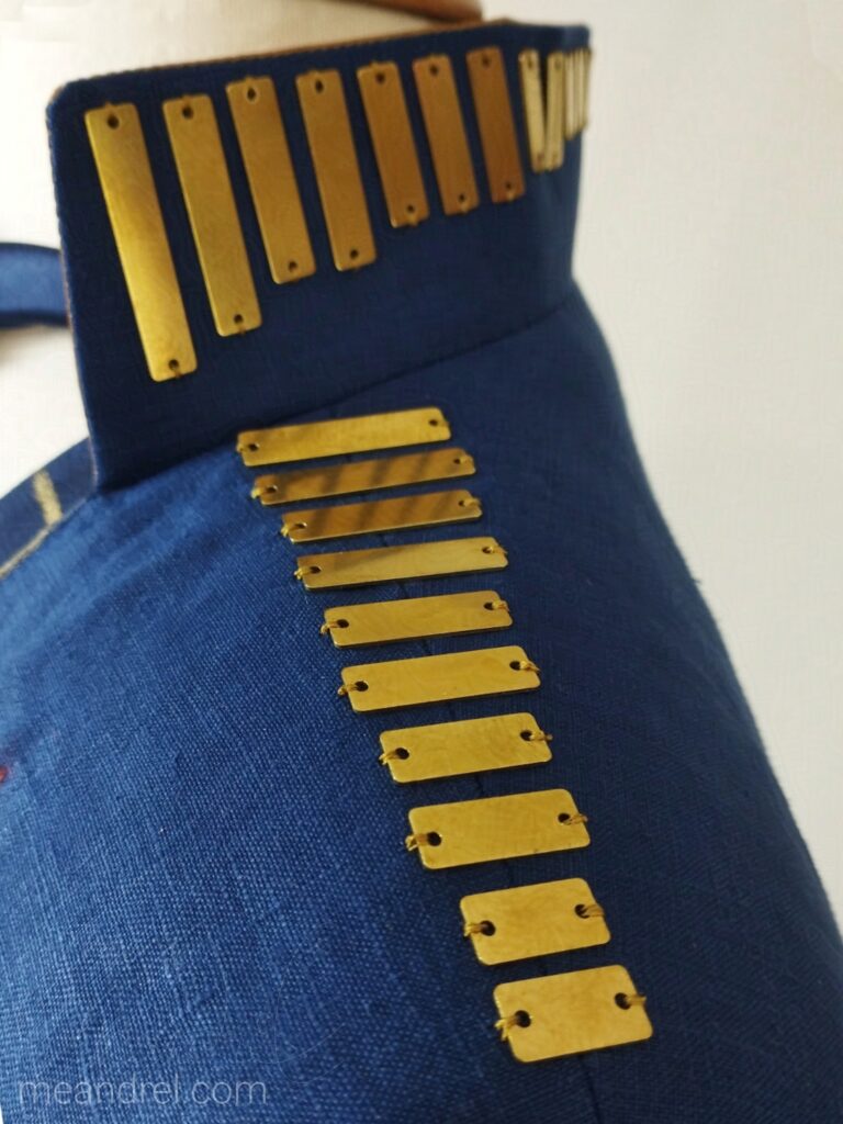 Photo de l'épaule et du côté du col, où sont cousues en ligne des plaques de laiton rectangulaires, de tailles dégradées.