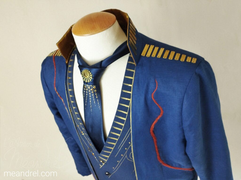 Photo prise en contre-plongée, du haut du costume sur mannequin : on voit bien les barrettes en laiton du col et des épaules, la cravate décorée et le haut du gilet ; ainsi qu'un bout de la liane qui remonte sur l'épaule gauche.