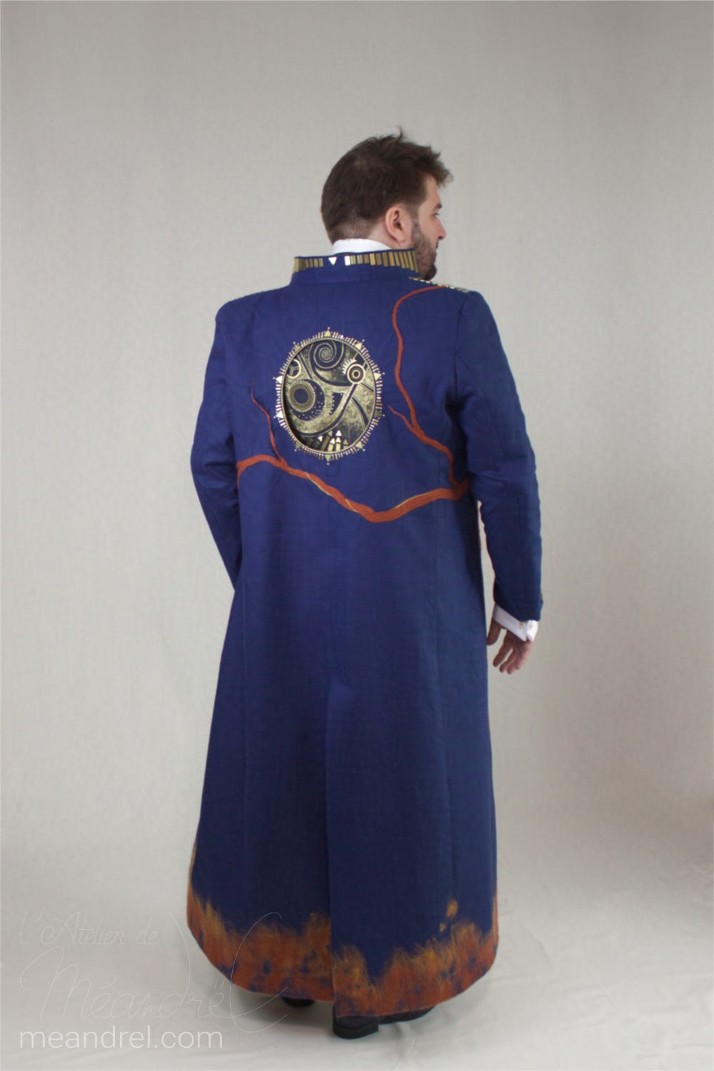 Photo de dos d'un long manteau bleu foncé, au bas nuancé de roux, et avec un cercle de motifs mécaniques dorés en haut.